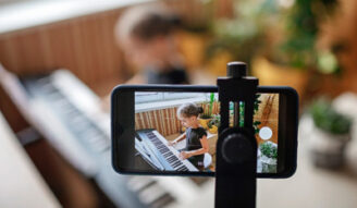 Pretty young musician in classic black dress playing digital piano at home during online concert and recording video, social distance during quarantine, online education concept, video blogging